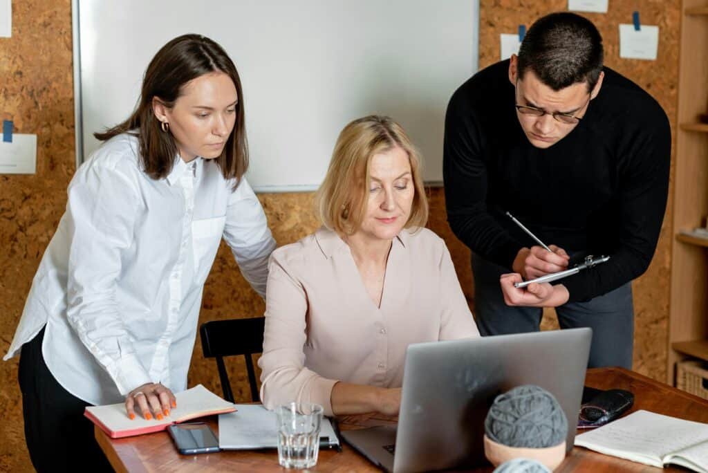 Two graduate students look over the shoulder of their research advisor who is demonstrating a process on a laptop.